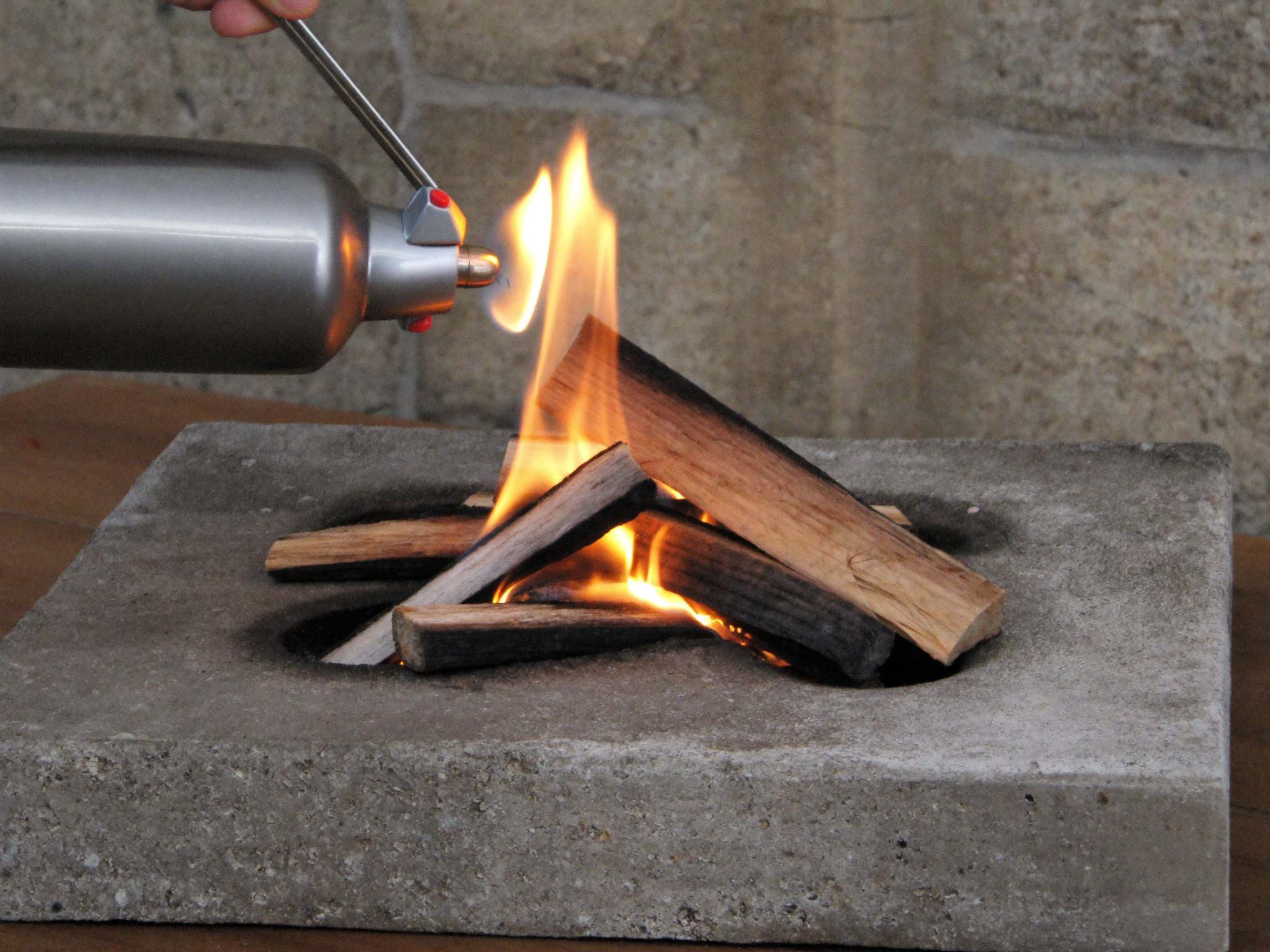 A hand turning the control knob on a propane fire pit table before ignition, with the fire glass bed visible around the burner.