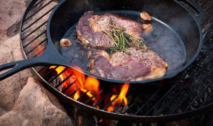 A seasoned cast iron skillet filled with sizzling steak and vegetables resting on a steel grate over a wood fire pit.