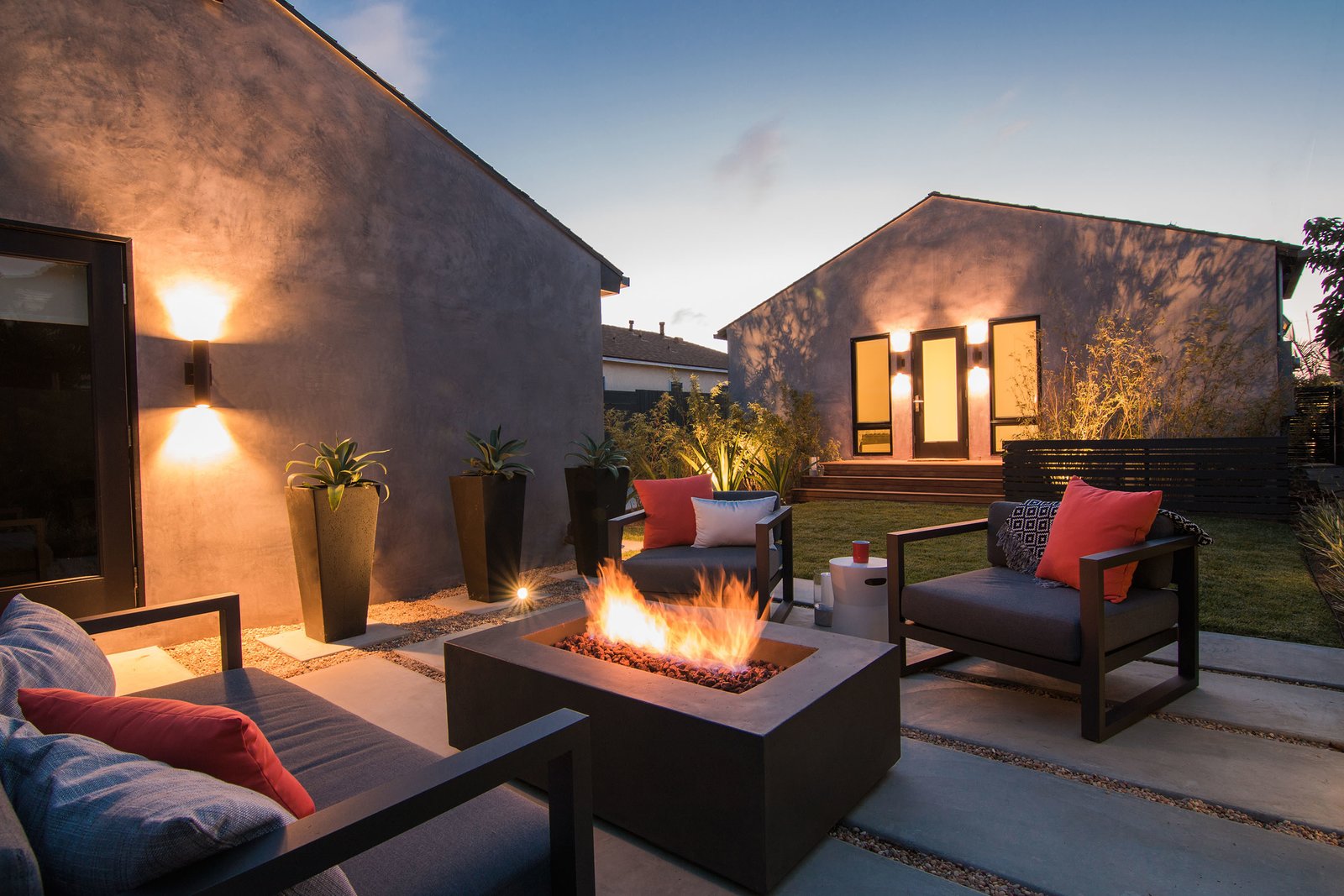 A cozy backyard patio setup at dusk, featuring a lit propane fire table surrounded by comfortable chairs and soft lighting.