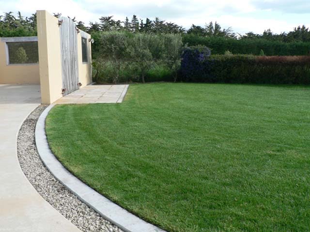 A shallow dirt trench dug through a green lawn, with a flexible yellow gas line laid inside, running toward a patio.