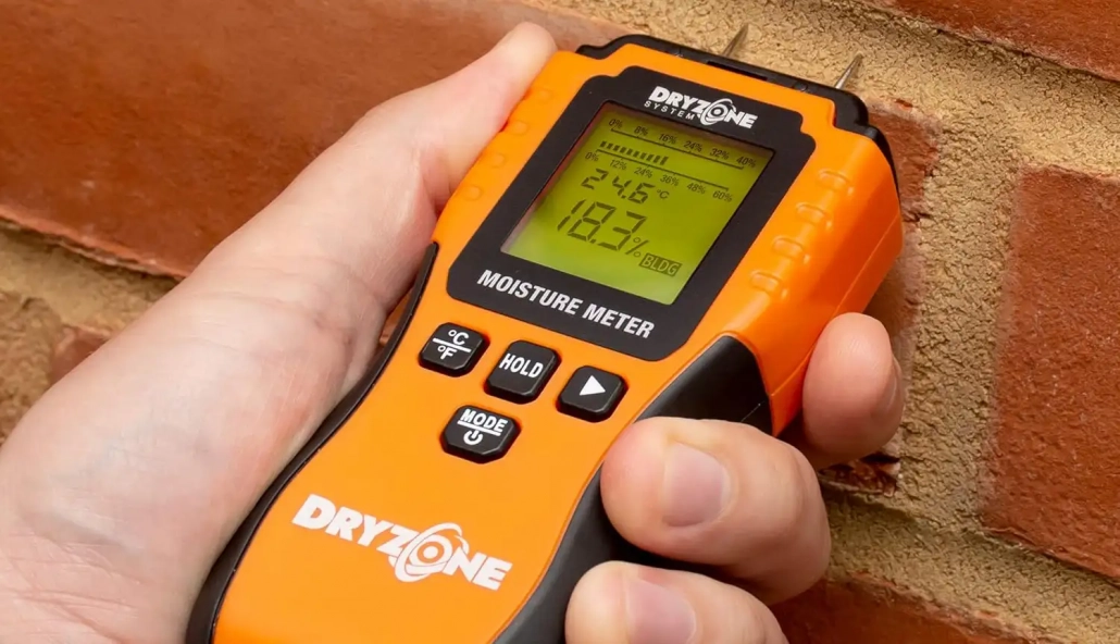 A hand pressing a digital moisture meter pins into a split oak log, displaying a reading below twenty percent.