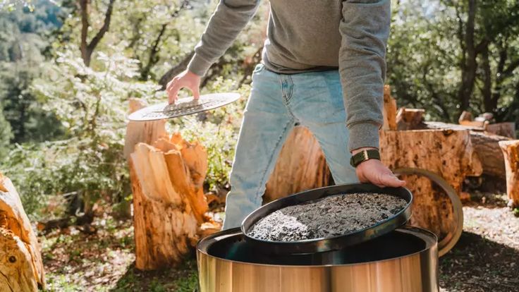 A hand using a small metal hand shovel to scoop grey wood ash out of a cold steel fire pit bowl into a metal bucket.