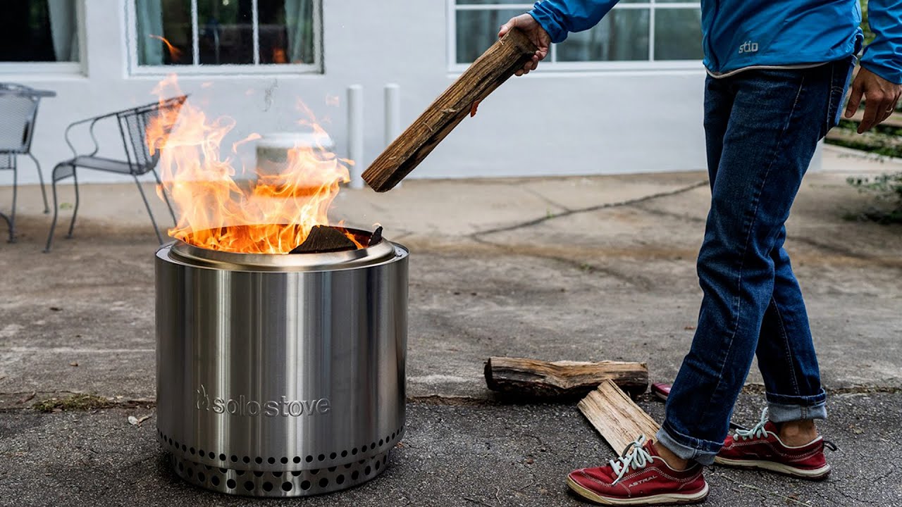 A stainless steel Solo Stove Bonfire fire pit burning dry firewood, with a bright secondary combustion flame emerging from the top ring vents on a concrete patio.